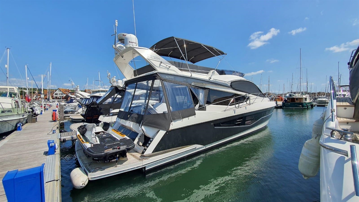 Luxury yacht docked at marina, featuring sleek design, large windows, and a jet ski on board, with boats and blue sky in background.